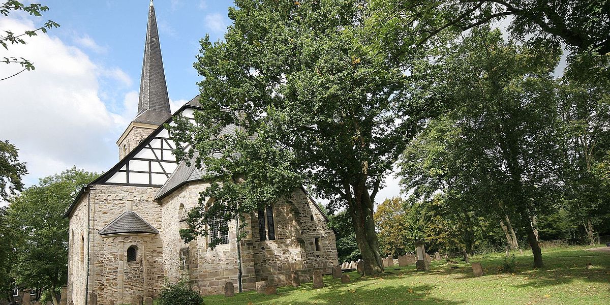 Dorfkirche in Bochum Stiepel von außen Eine historische Steinkirche mit einem hohen Kirchturm ist von großen Bäumen und einer Wiese umgeben, und um das Gebäude herum sind bei teilweise bewölktem Himmel Grabsteine zu sehen.