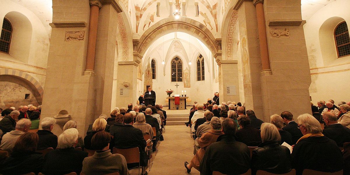 Festgottesdienst in der Dorfkirche Bochum-Stiepel Eine große Gruppe von Menschen sitzt einem Pfarrer gegenüber, der an der Stirnseite einer historischen Kirche mit hohen Gewölbedecken, Steinsäulen und sanfter, warmer Beleuchtung spricht.